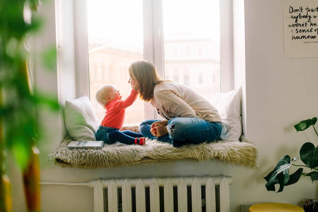 a mom with her toddler looking at her in front of a window touching the moms face